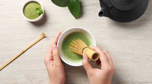 A vibrant green cup of matcha tea with a frothy top, whisked beside a bamboo whisk on a wooden table.